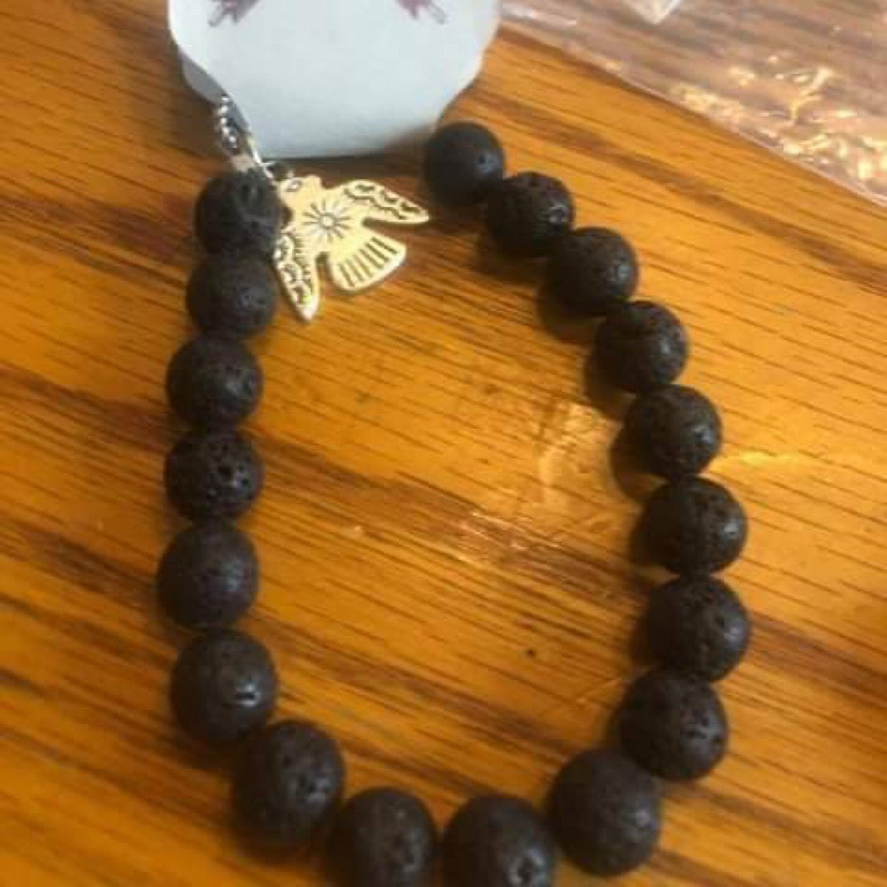 A black lava stone bracelet with a lobster claw clasp and a decorative flower-like charm, displayed on a wooden surface.